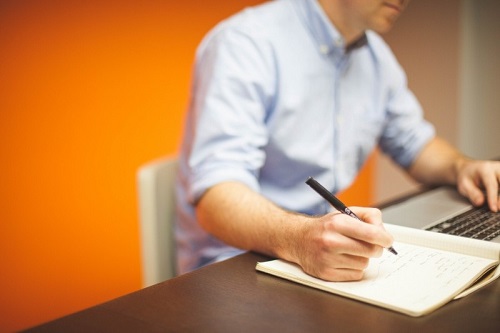 young-man-sitting-at-desk-using-laptop-and-writing-in-notebook.jpg